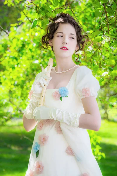 Happy woman musician in a beautiful long white dress decorated flowers with a flute in hand outdoors.