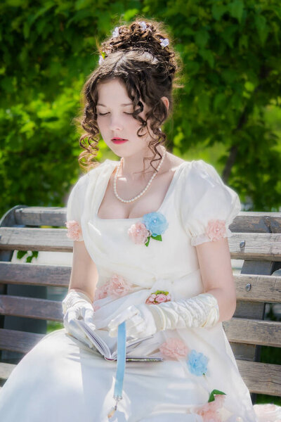 Portrait of a young bride woman in a historical white dress with a book in hands outdoors in a park on a bench.