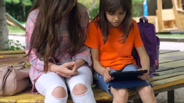 两个女孩正用平板电脑在户外坐在公园里 年轻的惊人的女孩看着电子小工具的屏幕 — 图库视频影像