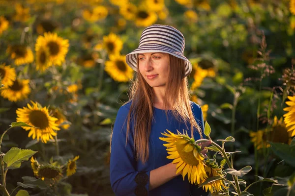 Jenta i blå kjole går med hatt i solsikkeåkeren. – stockfoto