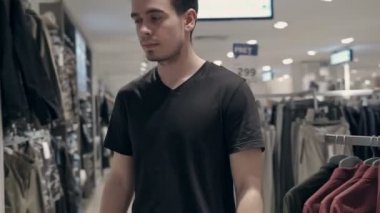 Genç adam bir mağazada kıyafet arıyor. Kumaşına dokunuyor ve satış alanındaki fiyatlara bakıyor..