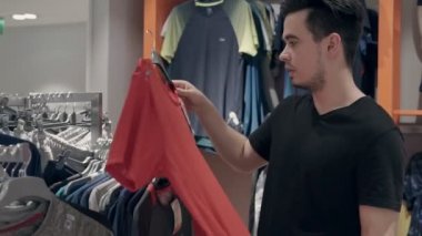 Genç adam bir mağazada kıyafet arıyor. Kumaşına dokunuyor ve satış alanındaki fiyatlara bakıyor..