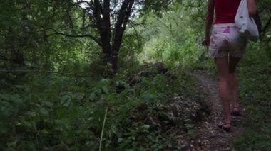 bluz ve kot şort arka görünüm kız yeşil orman boyunca yol boyunca yürüyor. Yavaş hareketler.