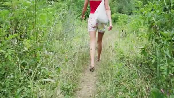 穿着上衣和牛仔短裤的背景女孩沿着绿色森林的小路行走 慢动作 — 图库视频影像