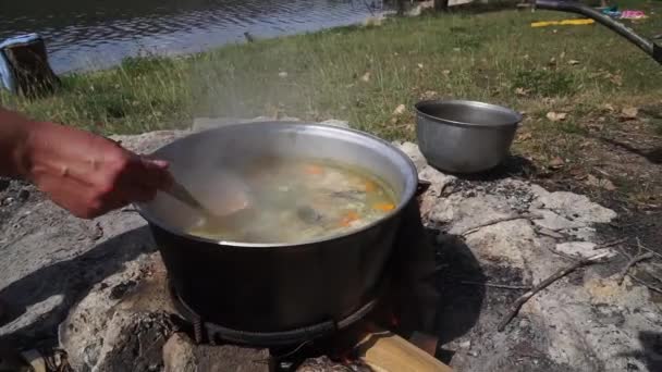 鍋を持つ手は 沸騰スープの材料を混合します クローズ アップ 自然の中で大きな鍋で調理 ハイキング旅行者のための食べ物 キャンプの観光客 — ストック動画