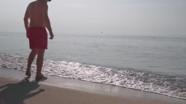 Kırmızı şortlu bir adam sahilde denizin kenarında duruyor. Güzel yaz sabahı.