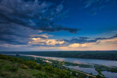 Yeşil manzaralı bir nehir ve mavi gökyüzü ile dramatik bulutlar.