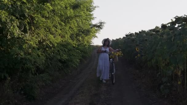 Strzał młodej kobiety w lekkiej sukience i kapeluszu jeżdżącej na rowerze po wiejskich drogach — Wideo stockowe