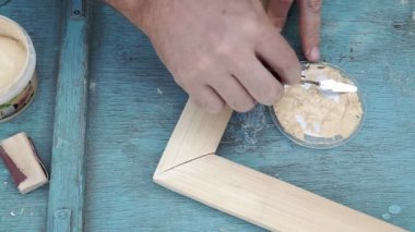 Usta çivi başlığını açık gri kaplamayla masa yüzeyine koyuyor. Marangoz masanın köşesini kapamak için sağ eliyle palet bıçağı kullanır ve daha fazla spatula kullanır.