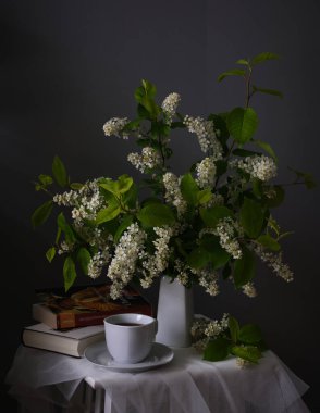 Sakin bir yaşam kompozisyonu açık bir kitap, narin bir çay fincanı ve canlı, gür bir çiçek aranjmanı sergiliyor.