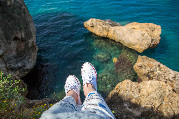 Молодая девушка в ветреном платье и кроссовках сидит на скале над морем
