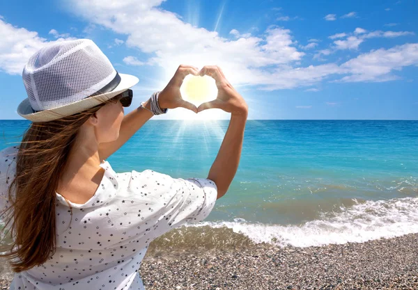 Brunette young girl holding hands in heart shape framing setting sun on sea beach