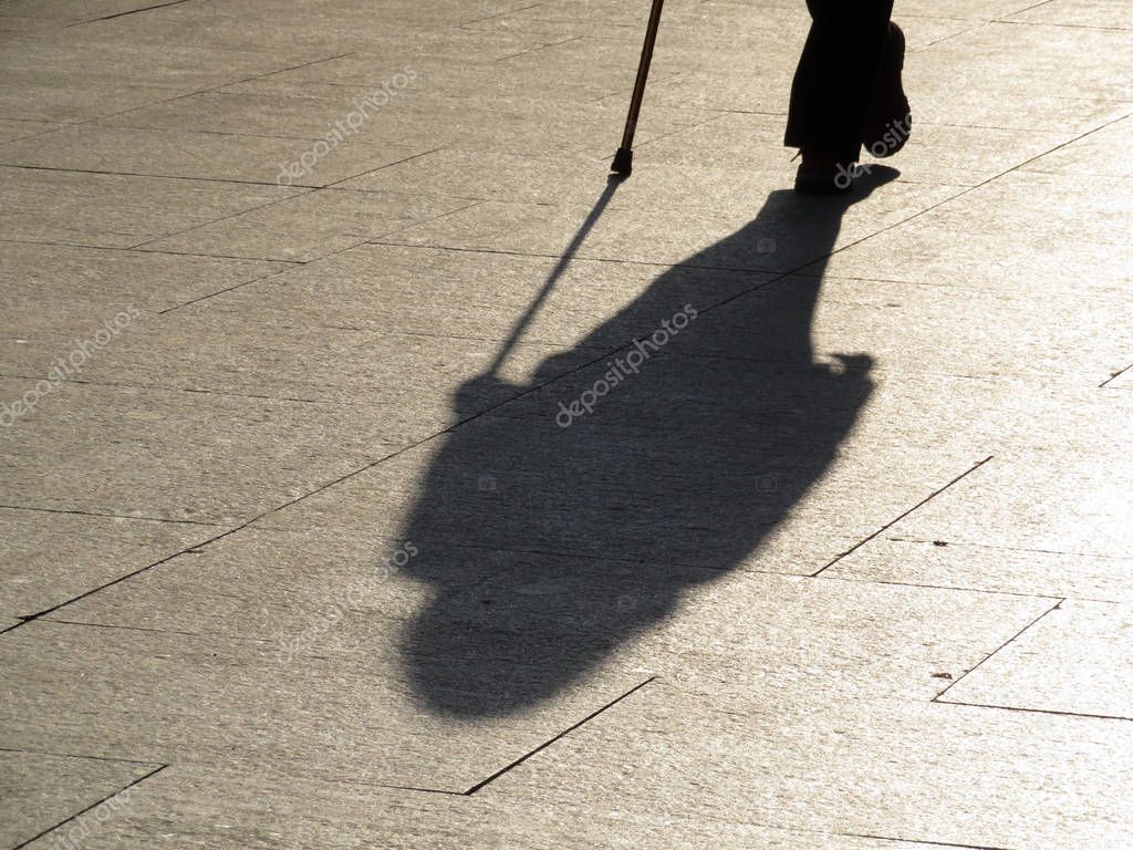 Person walking with a cane, black silhouette and long shadow on pavement. Concept of old age, blind man, limited ability, senior adult, dramatic life