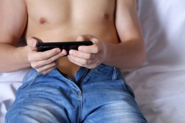 Smartphone adam çıplak gövde yatakta rahatlatıcı ile sıkıştırması kot pantolon erkek elinde. Kavram izlerken video, mobil oyunlar evde oynarken, seks dating, online iletişim, görüntülü sohbet