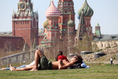 Moscow, Russia - April 2019: People sunbathing on a green grass in the Zaryadye park on background of the Kremlin towers and St. Basil's Cathedral. Hot weather in Moscow, russian spring leisure clipart