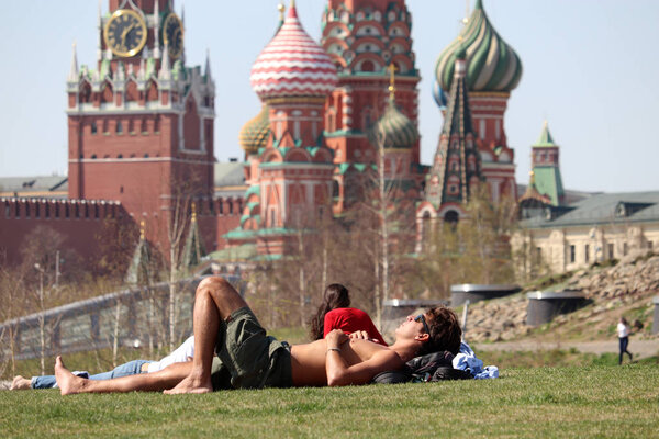 Moscow, Russia - April 2019: People sunbathing on a green grass in the Zaryadye park on background of the Kremlin towers and St. Basil's Cathedral. Hot weather in Moscow, russian spring leisure