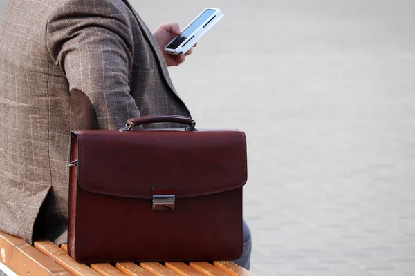 Man in a business suit sitting with a leather briefcase on a bench and using smartphone. Concept for businessman, official, government employee