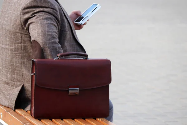 Bir bankta deri bir evrak çantası ile oturan ve akıllı telefon kullanarak bir iş elbisesi içinde adam. İşadamı, memur, devlet memuru kavramı