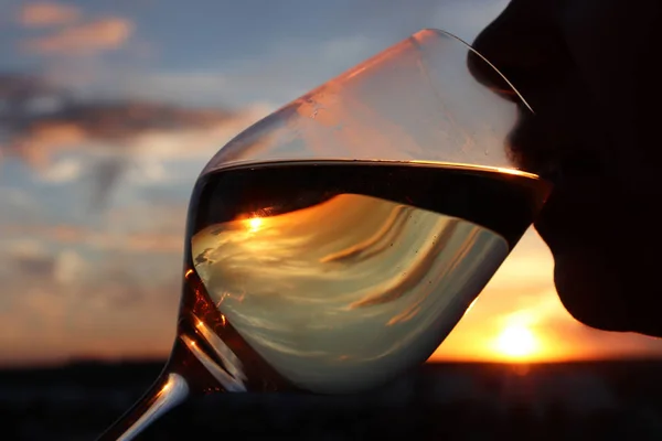 Silhouette of woman with glass of white wine at sunset, colorful sky is reflected in the drink. Girl drinking alcohol, romantic dinner at the resort, luxury life