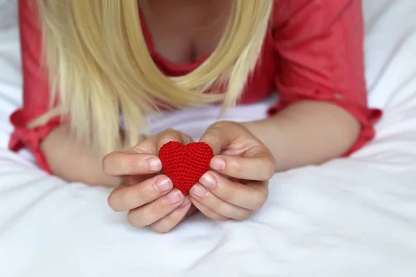 Woman in love, red knitted heart in female hands. Blonde girl lying on the bed, concept of romantic date, health care, innocence or organ transplantation