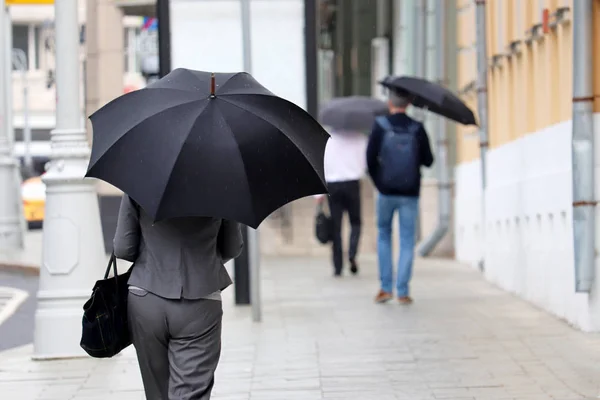 Rain in a city, slim woman with black umbrella walks on a street, rear view. People in rainy weather, summer storm