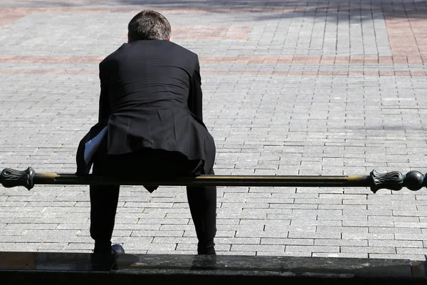 Man in a black business suit sitting on a railing on a city street, rear view. Concept of businessman, dismissed office worker, official, government employee