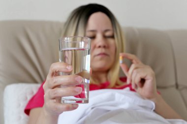 Elinde su bardağı olan bir kadın yatakta hap alıyor. Kapsül de ilaç, antibiyotik kavramı, vitamin, soğuk algınlığı ve grip