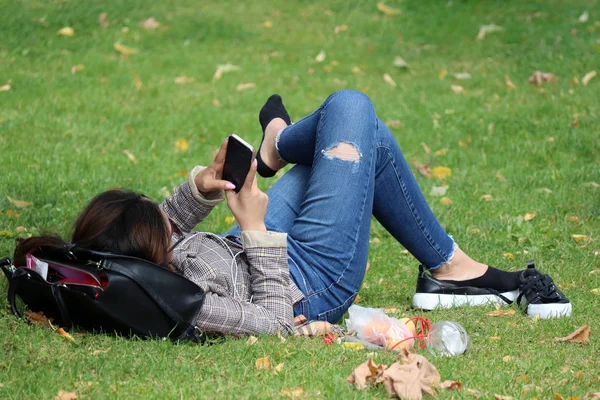 Girl with smartphone lying on a green grass with her shoes off. Young woman in ragged jeans relaxed on summer meadow, concept of tired legs, lunch break, online addiction