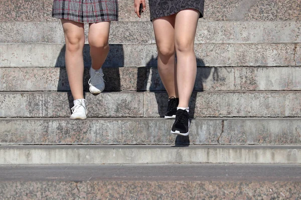 在阳光明媚的日子里 两个穿着短裙的苗条女孩沿着城市街道的台阶走下来 运动鞋 青年时装 服装和鞋类的女腿 — 图库照片