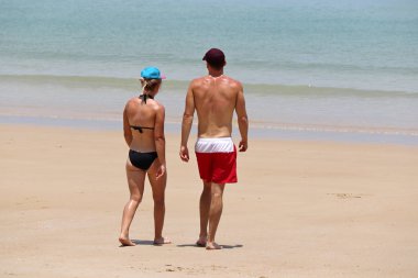 Çift tropikal mavi denizde yüzmeye gidiyor, renkli bir manzara. Siyah bikinili seksi bir kız ve kırmızı şortlu bir adam kumsalda yürüyorlar, romantik bir eğlence ve plaj tatili