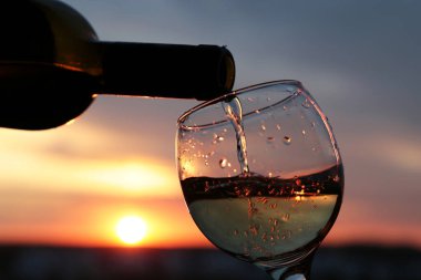 Güzel günbatımı arka planında beyaz şarap şişeden cama akıyor, turuncu güneş bulutların arasından parlıyor. Kutlama anlayışı, romantik akşam yemeği.