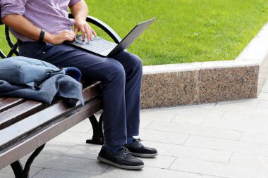 Yeşil çimenlik arka planda bir yaz şehrinde dizüstü bilgisayarlı bir adam oturuyor. Klavyede erkek eli, iş adamı konsepti, açık havada çalışma, serbest çalışma