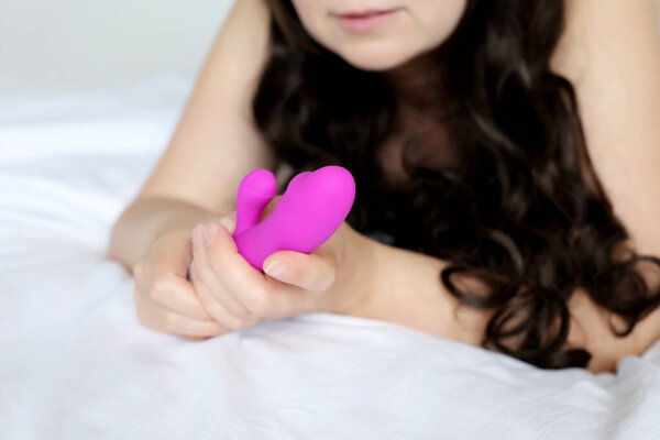 Vibrator in female hands, sex toys concept. Woman with long curly hair laying on the bed and holding purple silicone G-spot dildo, masturbation device