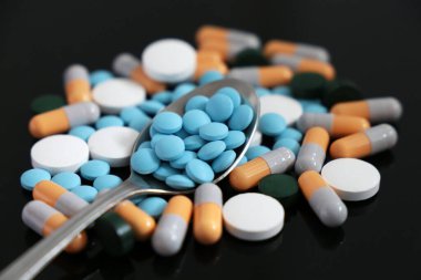 Haplar ve kapsüller, farklı ilaçlarla dolu kaşık ve siyah cam masaya saçılmış tabletler. Eczane, antibiyotik, vitamin kavramı