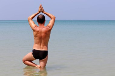 Yoga on the beach, man stands in a tree pose in the blue water of tropical sea. Balance with nature, fitness at sunny day, relaxation and exercise at vacation