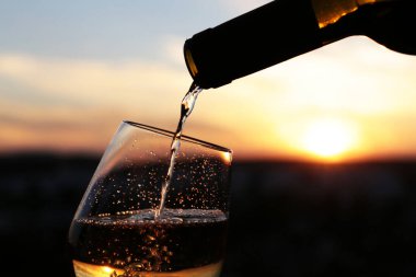Beyaz şarap, güzel bir günbatımı arka planında bardağa dökülüyor. Seçici bir odak noktası. Kutlama anlayışı, romantik akşam yemeği.