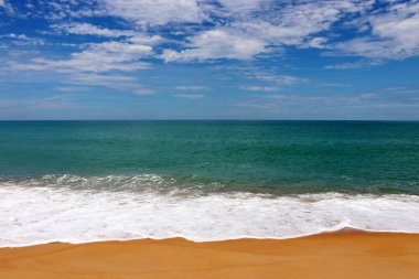 Tropikal bir kumsalda sörf, boş deniz manzarası, sarı kum ve beyaz köpüklü zümrüt dalgası. Mavi gökyüzü ve beyaz bulutlarla resimli deniz manzarası