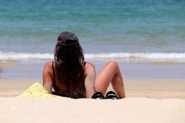 Sörf arka planında bir yaz plajında oturan bikinili kadın, bronzlaşmış deri kumla kaplı. Deniz tatilinin tadını çıkarma kavramı