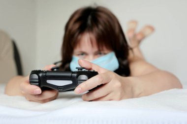 Medikal maske takmış bir kız, yatakta elinde joystick ile oyun oynuyor. Oyun sahası kadın ellerinde, kadın oyuncu koronavirüs salgını sırasında karantinada.