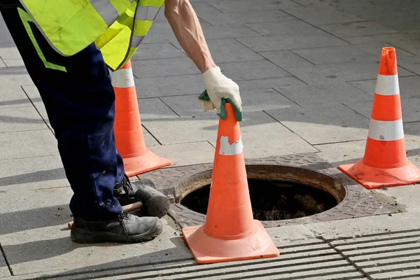 Trafik konilerinin yakınındaki bir sokakta açık kanalizasyon kapağının üzerinde çalışan bir işçi. Kanalizasyon, yeraltı tesisleri, su tedarik sistemi, kablo döşeme, su borusu kazası.