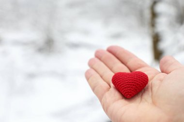 Kar ormanına karşı bir erkeğin elinde kırmızı örülmüş kalp. Romantik aşk kavramı, Noel tatili, Sevgililer Günü, kış havası veya hayırseverlik.
