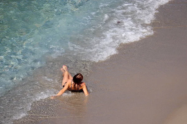 Beach vacation, relax and leisure by the sea. Girl in bikini and coral flip flops sunbathing on a beach lying on sand in surf waves, aerial view