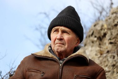 Deri ceketli yaşlı bir adam portresi ve mavi gökyüzü arka planında dağlarda örülmüş bir şapka. Sert dağcılık, seyahat ve özgürlük, sağlıklı yaşlılık ve doğa macerası kavramı