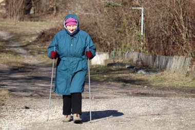 Kırsal yolda çubuklarla yürüyen yaşlı bir kadın, sağlıklı omurga ve eklemler için spor egzersizleri. Soğuk havada İskandinav yürüyüşü, sağlık tedavisi