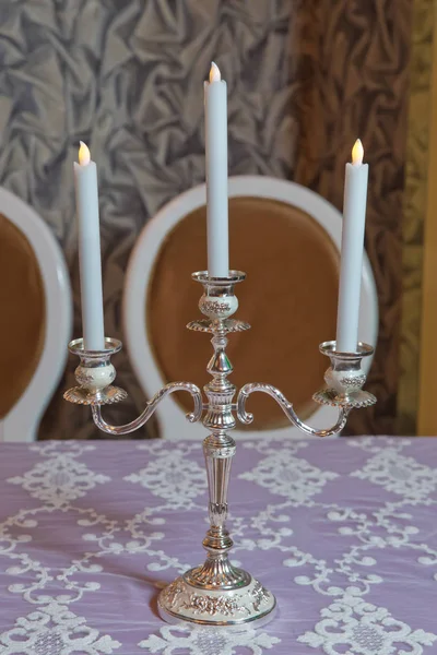 Three arms candle stick holder, two candelabra atop, on top of furniture, table against white wall in living room, house, home bedroom . Three white candles on the table .