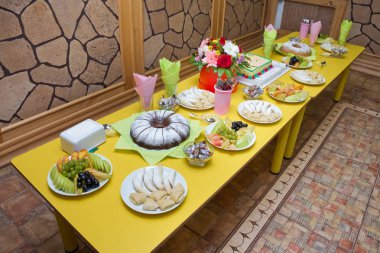 Sarı masa . Anaokulundaki çocuklar için tatil masası. pudra şekeri ile ev yapımı bundt kek. Elma turşusu ve armut turşusu. Tabakta siyah ve beyaz üzüm. Rendelenmiş muz tatlıları .