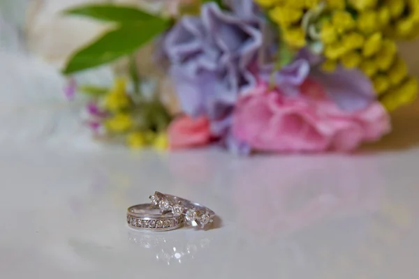 Bela imagem tonificada com anéis de noivado encontram-se em uma superfície de madeira contra o fundo de um buquê de flores. anéis de casamento e belo buquê como acessórios de noiva  . — Fotografia de Stock