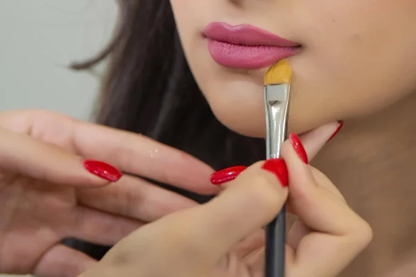 Güzel kadın yüzü. Makyaj ustasının eli, genç güzellik mankeni kızın dudaklarını boyama. Makyaj sanatçısı profesyonel bir fırça ile makyaj ve renkli ruj uygulamak. Makyaj sanatçısı kırmızı ruj uygular .