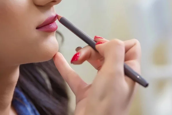 The female hand of visagiste is holding a pencil and drawing lips . Professional Makeup artist applies pencil lipstick bridal . Close up of unrecognizable pretty girl getting her lips painted.