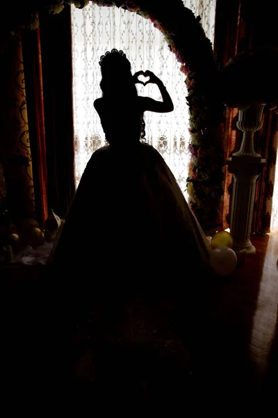 De bruid kijkt uit het enorme raam in de bruidssuite en onthult de achterkant van de trouwjurk. De achterkant van een witte bruiloft houdt een hart in zijn hand. — Stockfoto
